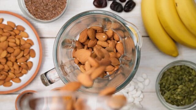 Top view of many whole almonds pouring into blender on table with various nuts and fruits. Preparing almond milk as base for plant-based vegan smoothie without animal fats. Healthy vegan drink.