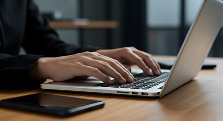 Close-up of hands typing on a laptop keyboard with a smartphone on a wooden desk, representing business and technology.