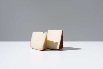 Dos trozos de queso de oveja curado y de cabra sobre una mesa blanca y fondo gris.