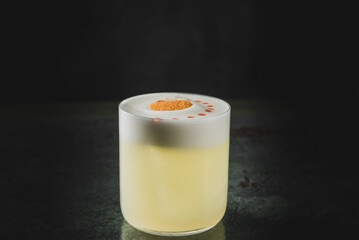 Elegant cocktail in a clear glass with white foam, orange garnish, and red dots, set against a dark...