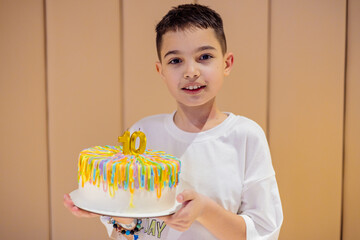 A young boy of Middle Eastern descent proudly holds a colorful birthday cake adorned with bright...
