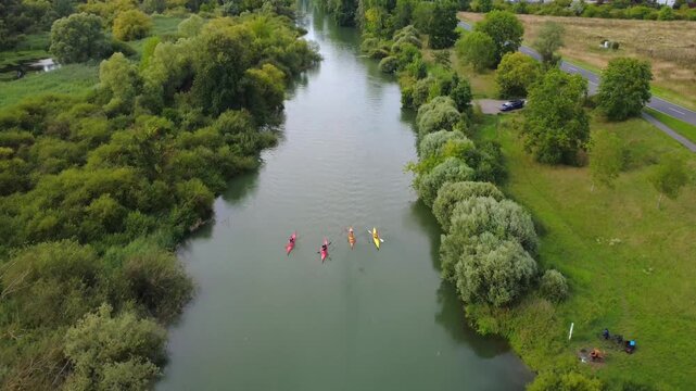 Aerial 4K drone footage of a peaceful river surrounded by lush green trees and bushes. Four colorful kayaks are paddling calmly downstream in a serene natural setting. Perfect for nature, outdoor spor