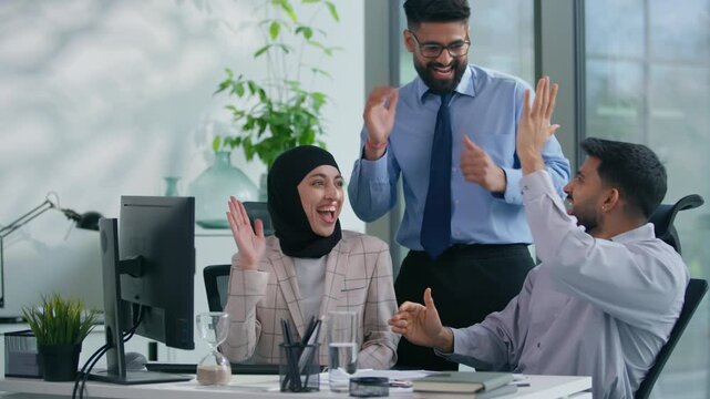 Excited Arabian Muslim business team celebrating success in office achieve goal looking at computer cheering smiling giving high five feeling happy teamwork motivation career partnership win teamwork