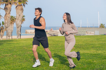Young fit couple jog side by side on a green seaside lawn, smiling at each other while maintaining...
