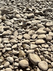 Close-up view of smooth, weathered river stones in various shapes and sizes, scattered naturally. The soft beige and grey tones highlight the organic texture and natural beauty of the rocky surface.