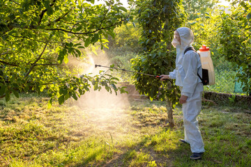 A gardener in a protective suit, mask and glasses uses a professional sprayer to treat the garden with insecticides and fungicides from pests