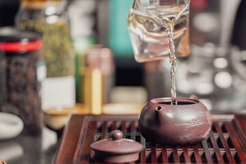 Close-up of traditional tea ceremony with clay teapot, glass pitcher pouring hot water, and tea accessories on a wooden tray in warm ambient light