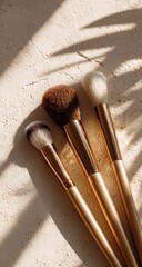 Four gold-handled makeup brushes with varied brush heads lie on a textured beige surface, partially shaded by a palm frond shadow