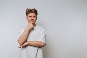 A young man in a white t-shirt stands against a plain background, looking thoughtful with his hand...