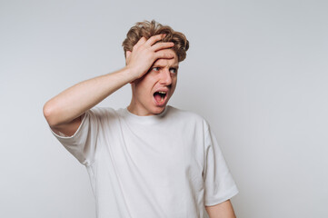 A young man in a white t-shirt looks surprised and holds his head with one hand against a plain...
