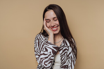 Fototapeta premium A woman with long dark hair smiles gently, resting her face on her hand. She wears a zebra-patterned shirt and a pearl necklace, standing against a beige background.