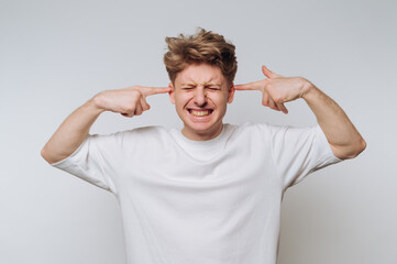 A young man in a white t-shirt covers his ears with his fingers, eyes closed, expressing...