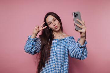 Young woman in blue checkered dress taking a selfie against a pink background.