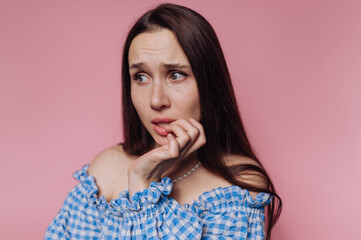 A worried woman with long dark hair biting her nails against a pink background.