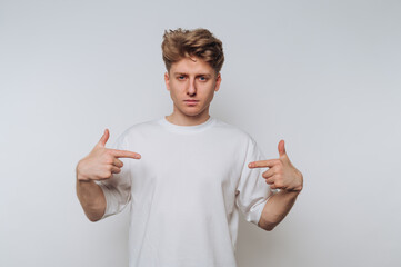 Young man in a white t-shirt pointing at himself against a plain background.