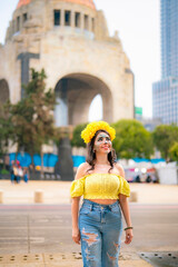 Fototapeta premium Smiling young Latina with colorful catrina makeup walks at Monumento a la Revolucion in Mexico City during Dia de Muertos, wearing yellow top and ripped blue jeans, radiating confidence and tradition