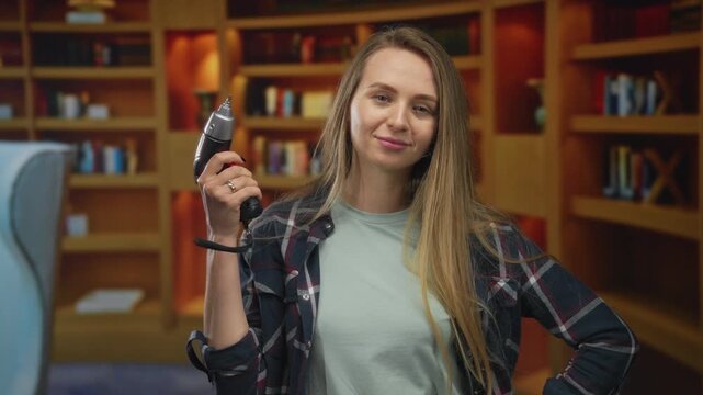 Woman smiling and holding a drill in a cozy library setting, showcasing a relaxed atmosphere with bookshelves in the background.