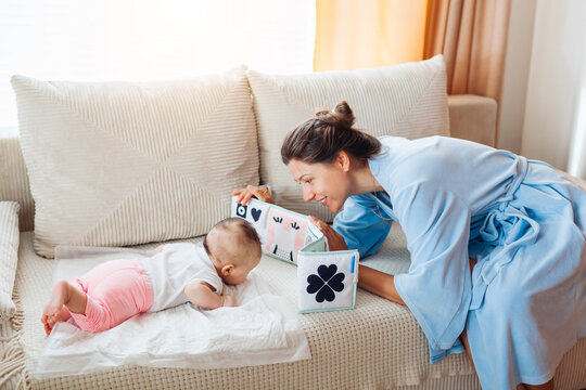 Young mother playing with newborn baby during tummy time at home. Infant looks at fold-out interactive high contrast book - Powered by Adobe