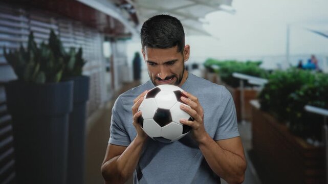 Young man outdoors in urban street holding soccer ball with a thoughtful expression, surrounded by urban elements under a canopy.