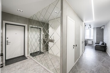 Modern apartment hallway with a large, geometric mirrored wall. Gray stone flooring and white doors add a sleek, minimalist touch