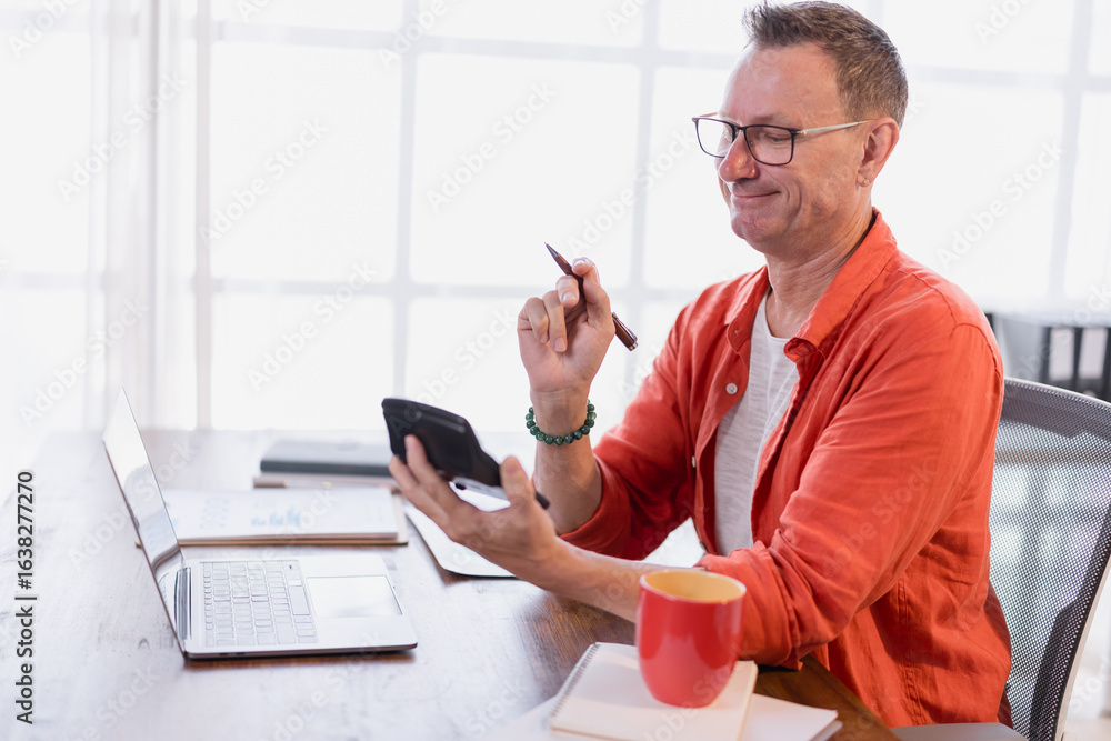 Poster smiling entrepreneur holding a pen and using a calculator while working on a laptop at a desk in a b
