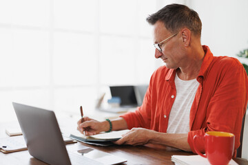 Obraz premium Mature businessman writing notes in his notebook while working remotely using his laptop, sitting at his desk in a bright home office, enjoying a cup of coffee