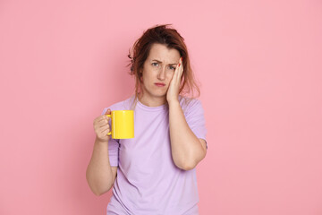 Woman with cup of coffee suffering from hangover on pink background