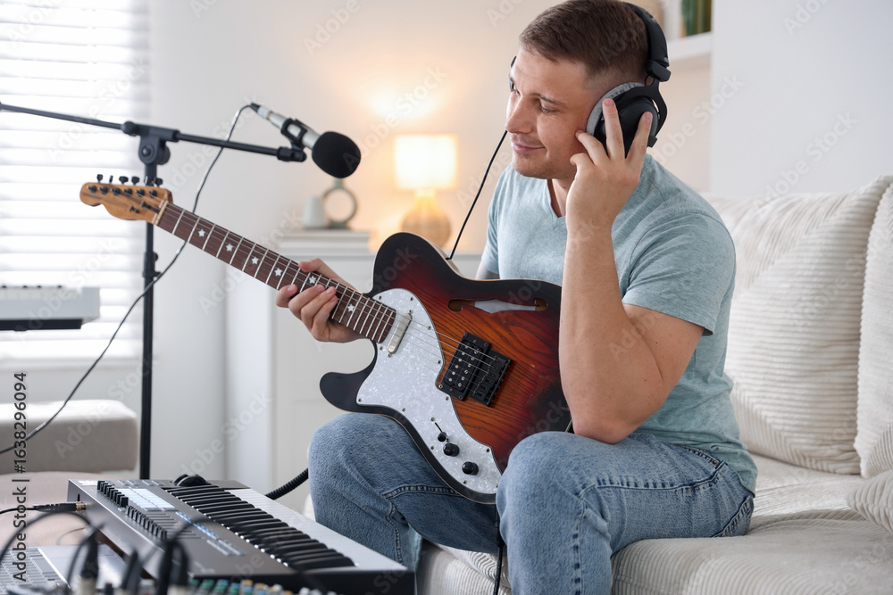 Poster man in headphones working with guitar and midi keyboard on sofa in home studio