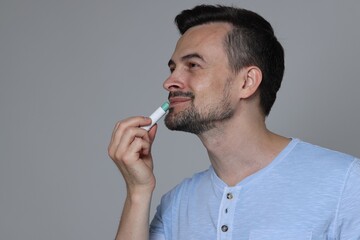 Handsome man applying lip balm onto his lips on grey background. Space for text