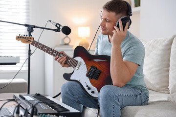 Man in headphones working with guitar and MIDI keyboard on sofa in home studio
