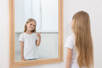 Beautiful little girl showing thumbs up while looking in mirror indoors