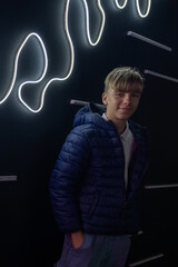 Portrait of smiling young blonde boy wearing blue puffer jacket against striking dark wall adorned...