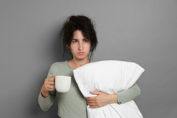 Woman with pillow and cup of drink suffering from hangover on grey background