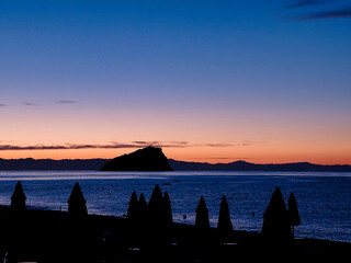 Umbrellas on a sandy beach at dawn as the sun peeks out between the umbrellas and the sea on a hot...