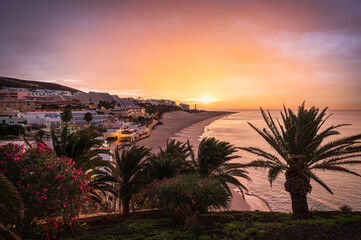 Morro Jable, Fuerteventura, at sunset with sandy beach, palm trees, and calm Atlantic Ocean under a colorful sky.