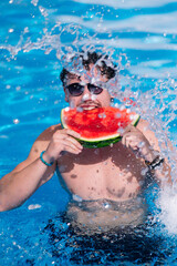 Young man eating juicy watermelon slice in refreshing swimming pool splashing water around playfully