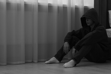 Sadness and depression. Man in hoodie near curtains at home, space for text. Black and white effect