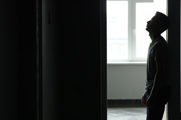 Fototapeta premium Sadness and depression. Tired man in dark room with open door against window with daylight. Space for text
