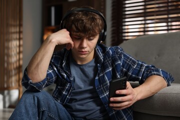 Teen problems. Upset teenage boy in headphones with smartphone near sofa at home