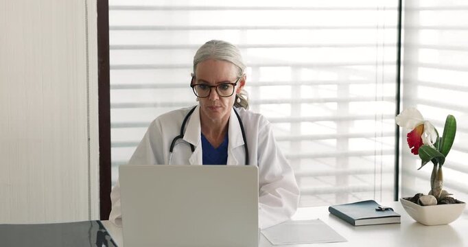 Mature 50s woman doctor sits at workplace desk, focused on laptop while takes notes on paper, updating patient medical records, writing symptoms, reviewing diagnoses, prescriptions or treatment plans