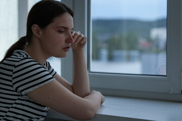 Sad young woman near window at home, space for text
