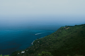 A serene view of a lush green headland stretching into the deep blue sea under a soft, hazy sky. The gentle gradient of ocean hues blends seamlessly with the horizon, creating a peaceful and minimalis