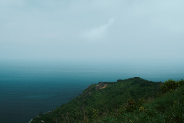 A serene view of a lush green headland stretching into the deep blue sea under a soft, hazy sky. The gentle gradient of ocean hues blends seamlessly with the horizon, creating a peaceful and minimalis