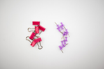 Pink binder clips and purple push pins arranged on a white background. Colorful stationery composition highlighting metallic textures and vibrant colors.