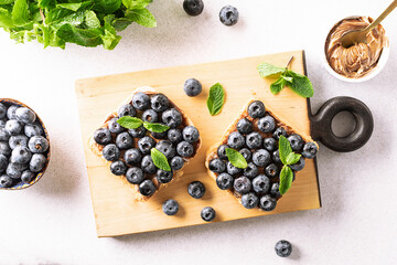 Sweet homemade toasts with chocolate paste, nuts, blueberry and aromatic mint leaves on the serving board on white stone background. Tasty summer dessert.