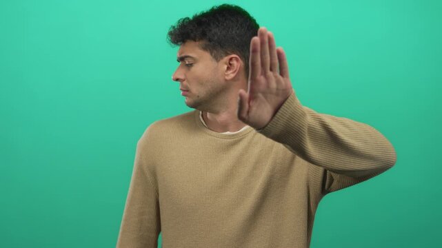Man with face expressionless covers eyes with hand against background where hispanic colored young guy in green sweater stands.