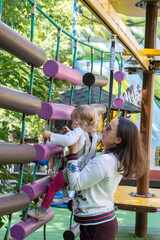 Fototapeta premium The mother supports her young daughter as she climbs on vibrant wooden beams at an adventure park. They enjoy a day filled with exploration, laughter, and outdoor activities.