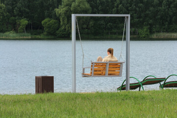 Young woman relaxing on swing near peaceful lake in daytime, rear view, serene summer but cool...