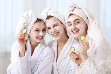 Happy women with facial masks and cucumber slices in bathrobes after spa procedures indoors