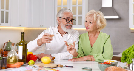 Elderly couple drinking white wine while cooking at table in kitchen